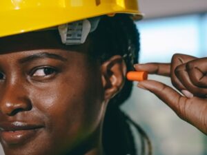 Person mit Schutzhelm setzt sich einen orangefarbenen Gehörschutzstöpsel ein